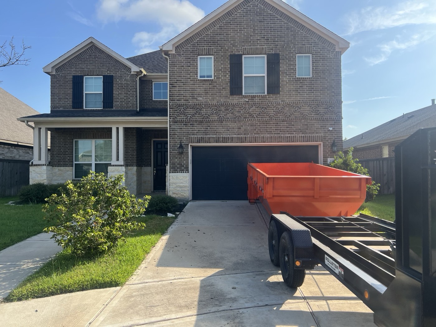 Orange 14-yard can on the roll-off trailer being delivered at a two-storey brick home, blue sky