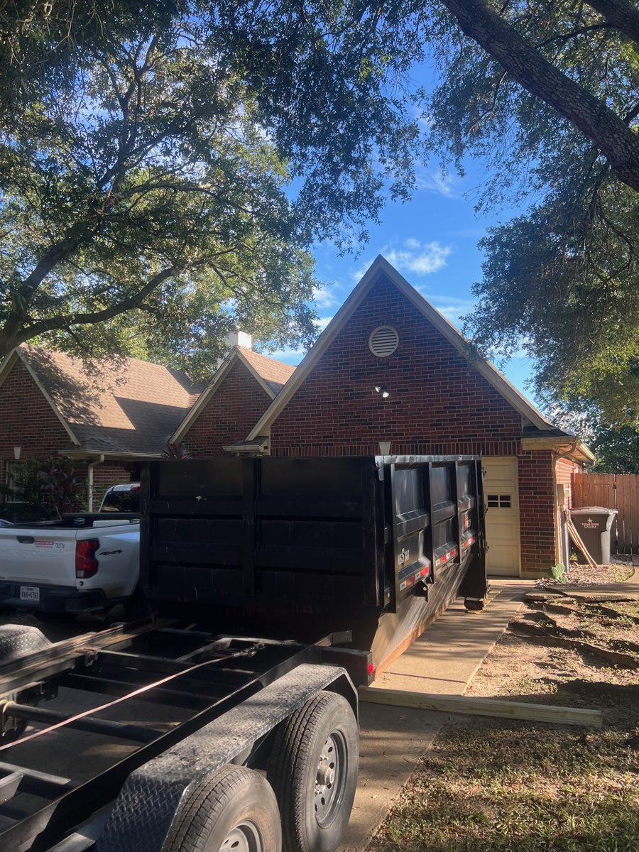 Roll-off dumpster set on the driveway of a brick home under oak trees